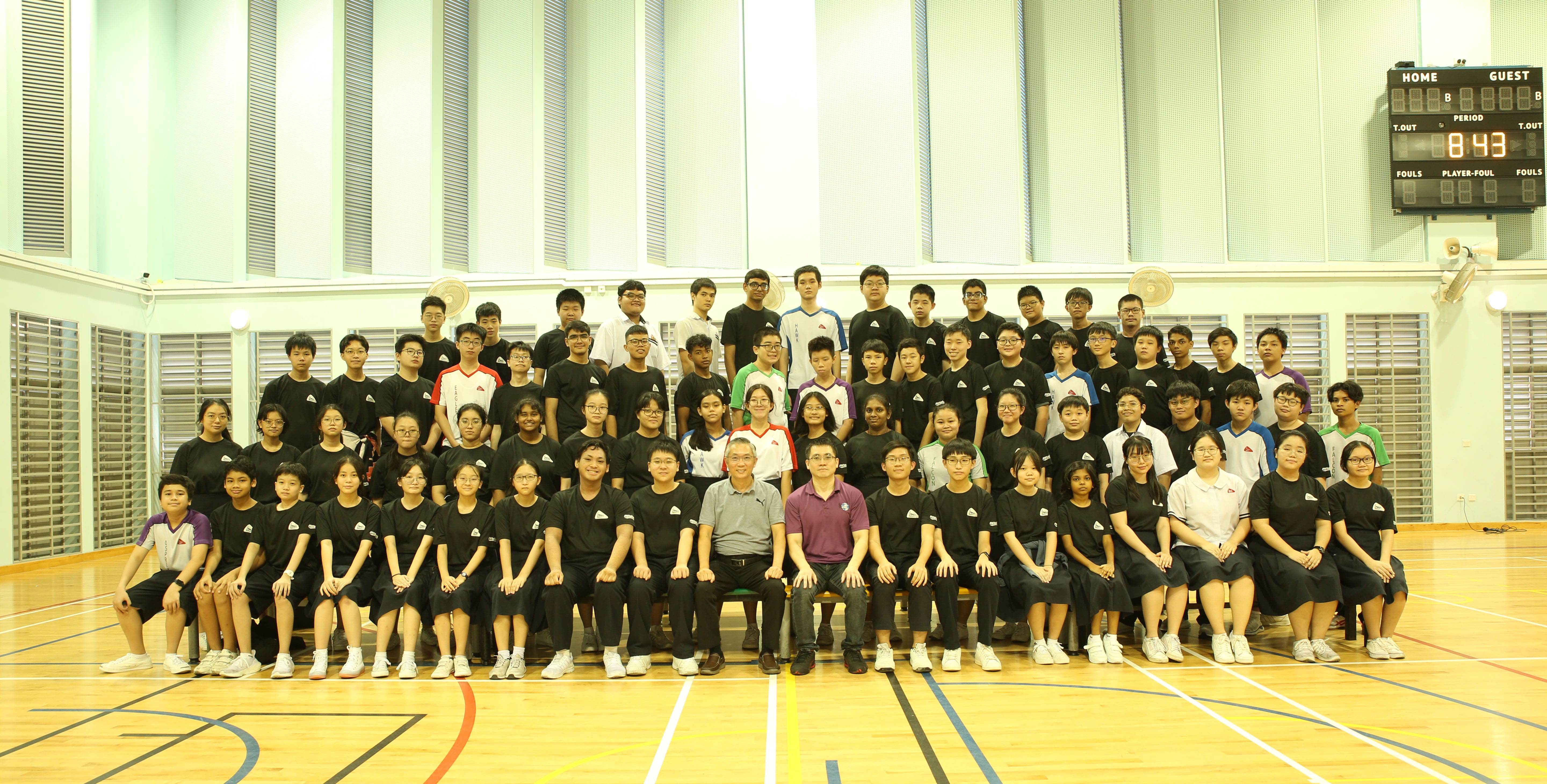 Large group of students in black uniforms posing with two adults in an indoor gymnasium with a scoreboard.